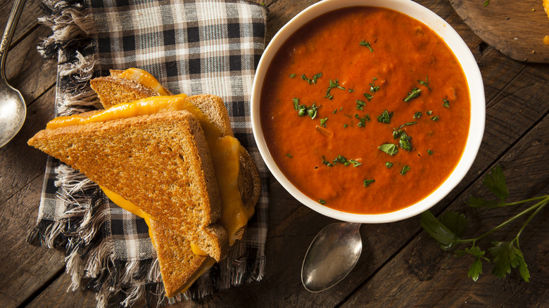 Top-down view of a grilled cheese sandwich on a plaid napkin next to a bowl of tomato soup