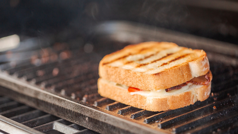 Grilling grilled cheese sandwich with bacon strips and fresh tomato on outdoor gas grill.