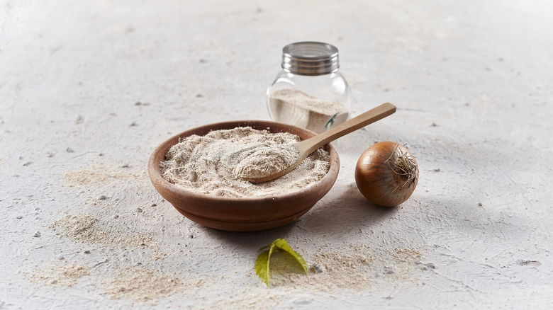 Wooden bowl of onion powder with a spoon next to a spice shaker and fresh whole onion