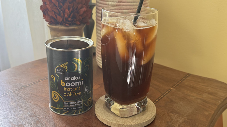 A glass of iced coffee sitting next to an open can of Araku Boomi instant coffee