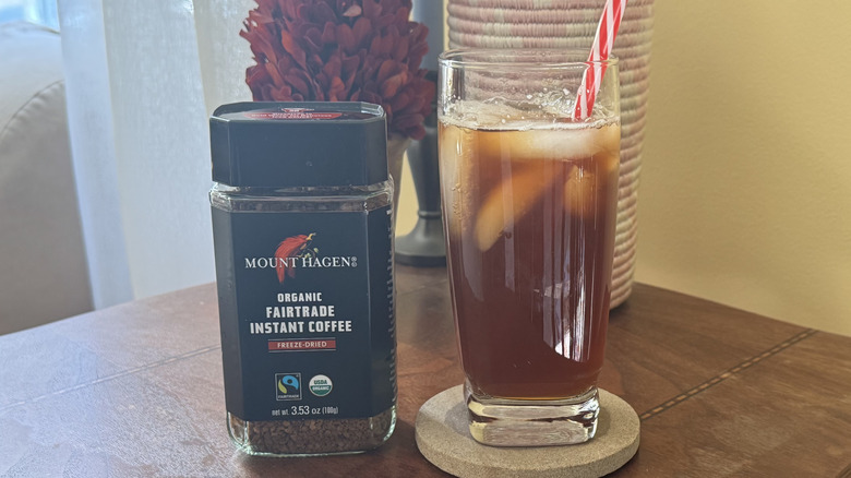 A glass of iced coffee on a table sitting next to a cannister of Mount Hagen Instant Coffee