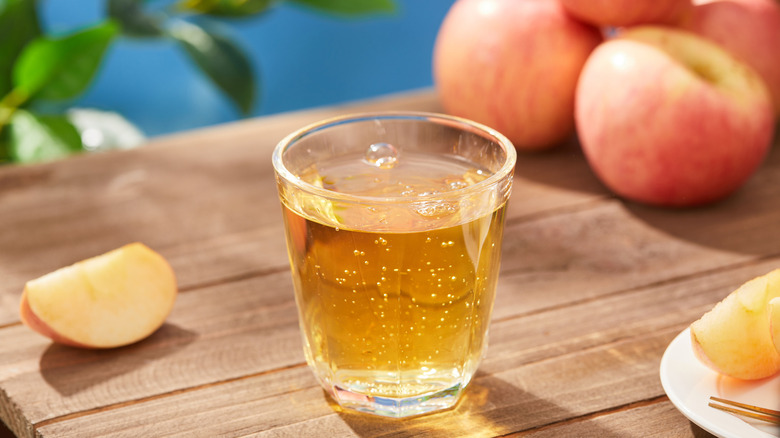 Apple juice in glass on wood cutting board in front of apples.