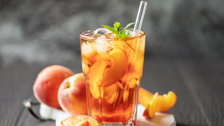 Peach iced tea in glass with straw in front of peaches.