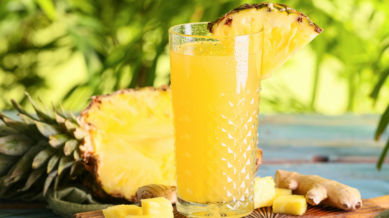 Glass of pineapple juice in a tall glass on chopping board in front of palm trees.