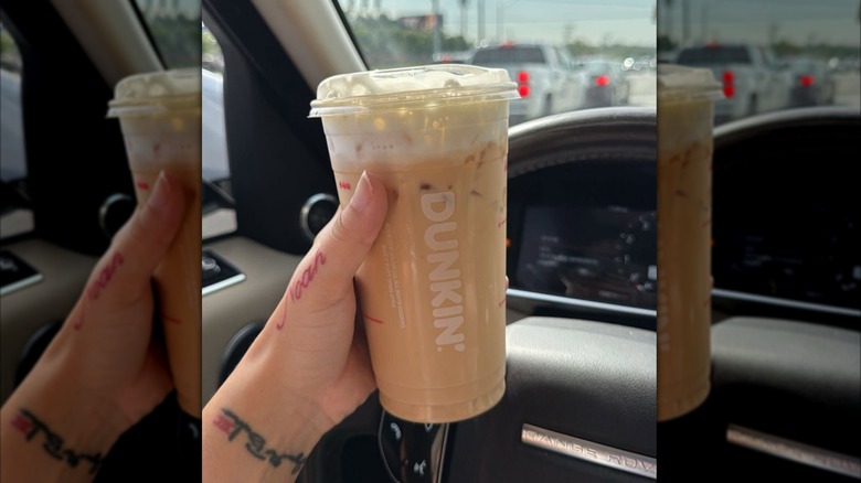 Woman's hand with tattoos holding an iced latte in a clear plastic Dunkin' cup in front of a steering wheel