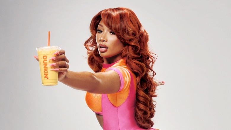 Megan Thee Stallion wearing pink and orange workout gear doing a lunge and holding a Megan's Mango Protein Refresher