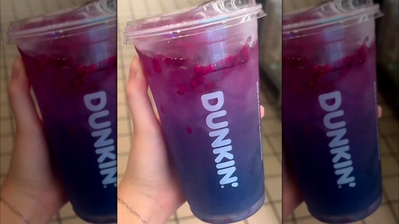 Hand holding a Starlight Lemonade, with a purple top layer and an indigo bottom layer with dragonfruit pieces, in a clear plastic Dunkin' cup