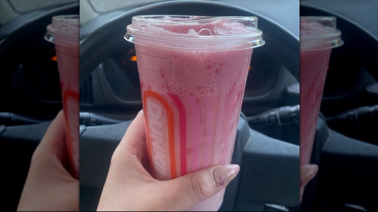 Woman's hand holding an Ultimate Pink Daydream Refresher in a plastic Dunkin' cup in front of a steering wheel