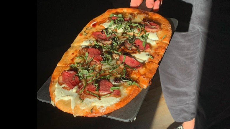 Oval-shaped Duck Bacon Flatbread Pizza held on a stone tray by a person in gray pants