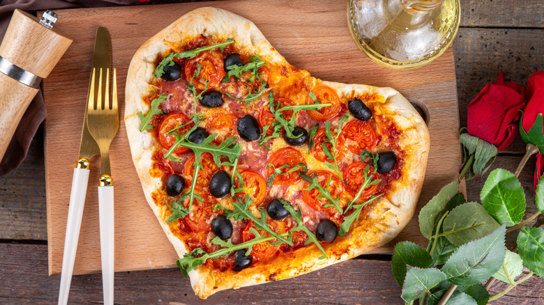 Heart-shaped pizza with red sauce, tomatoes, olives, and radicchio on a cutting board, with an olive oil carafe and white/gold utensils, two red roses to the side