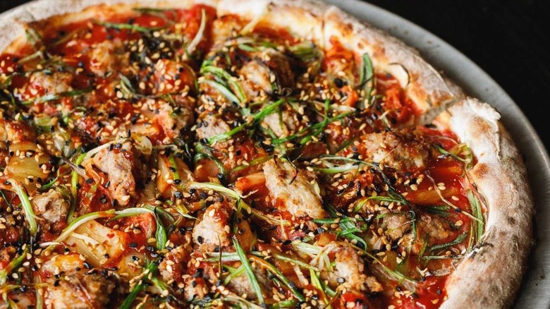 Partial view of Lady ZaZa pizza on plate, with red sauce, kimchi, greens, against a dark background