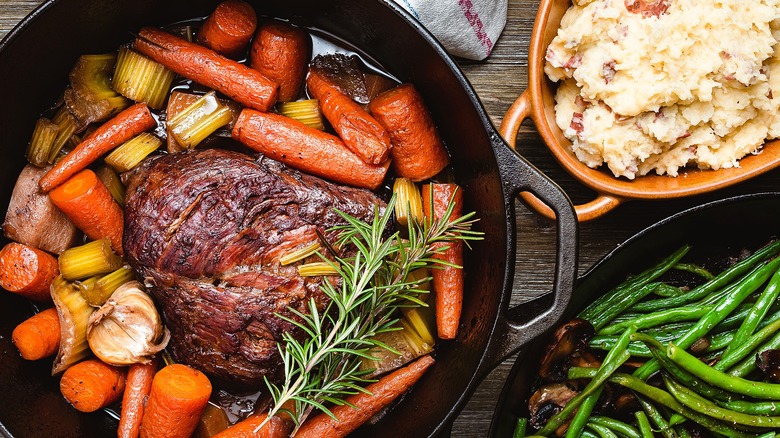 Pot roast and chopped vegetables in a cast-iron pan next to platters of green beans and mashed potatoes