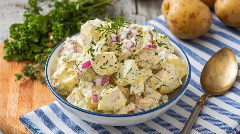 Creamy potato salad with fresh herbs and red onion in a white bowl over a blue-and-white-striped dish cloth