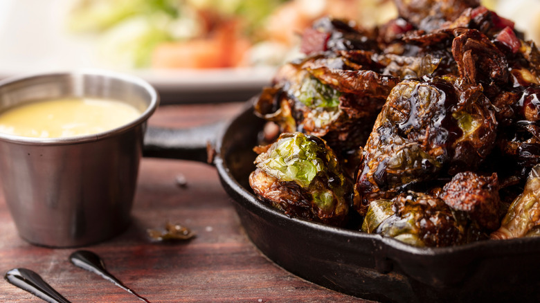 Balsamic glaze brussel sprouts in skillet on wood cutting board next to sauce.