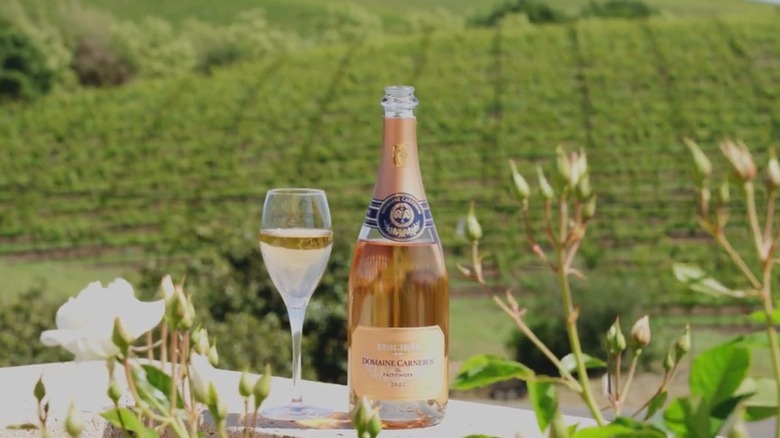 Bottle of Domaine Carneros next to a class in front of a winery backdrop.