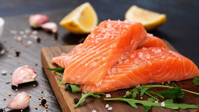 Two pieces of raw salmon topped with coarse salt on a wooden cutting board with fresh arugula,