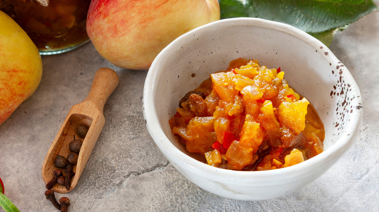 A white bowl of apple chutney on a grey table with apples and spices nearby
