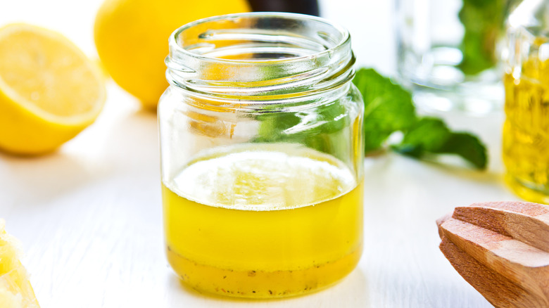 A glass container of citrus vinaigrette with lemons and herbs in the background