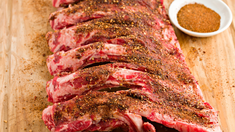 a rack of ribs covered in dry rub seasoning on a wooden board alongside a small bowl of seasoning