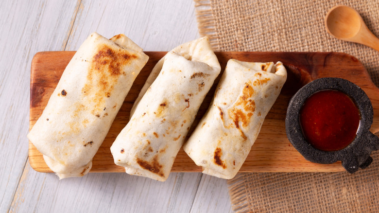 Three burritos on a wooden tray next to a bowl of salsa