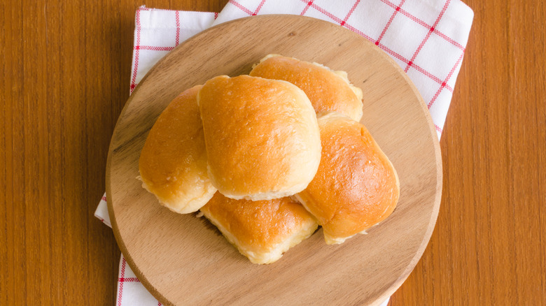 Dinner rolls piled on a wooden plate on top of red and white checkered cloth on table.