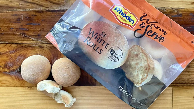 A package of Rhodes Warm & Serve Soft White Rolls beside two rolls outside the package on wood surface.