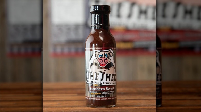 A bottle of the The Shed Original Southern Sweet BBQ sauce on a wooden table with The Shed logo on the wall in the background