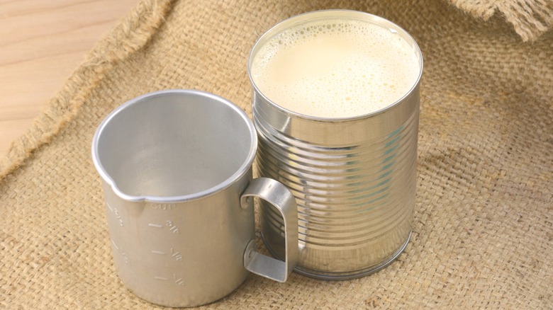 Open can of evaporated milk next to metal measuring cup.