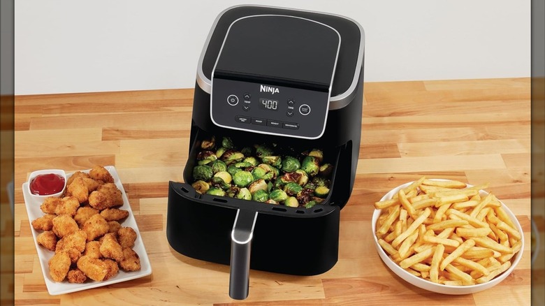 A black Ninja air fryer with brussels sprouts inside sitting on a table next to fries and nuggets