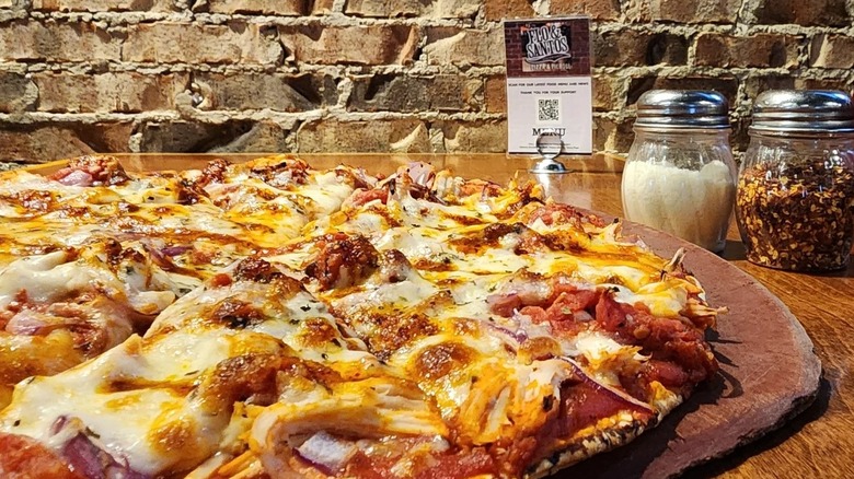 Flo & Santos loaded tavern-style pizza on table with parm and red pepper, brick background