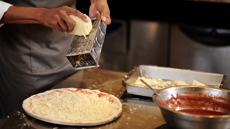 Chef grating cheese over a pizza with sauce and pan on counter