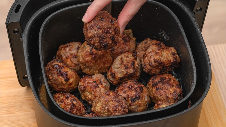 Fingers holding a meatball over an air fryer basket full of meatballs.