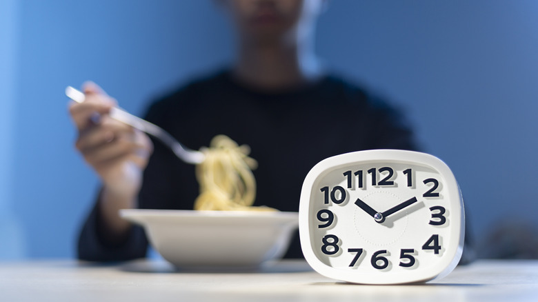 A clock in front of a person eating a bowl of noodles.