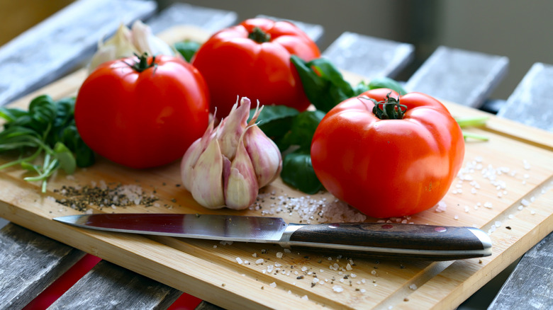 Garlic bulb and whole tomatoes with basil leaves on a cutting board with knife.