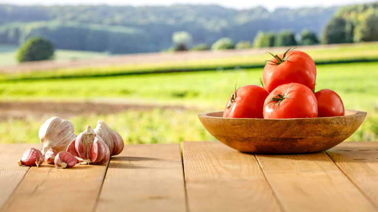Garlic bulbs and cloves and tomatoes in a wood bowl on a wooden table outdoors.