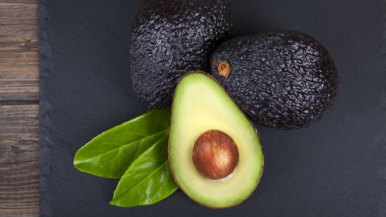 Two whole avocados and one avocado cut in half on a black cutting board