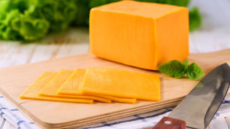 A block of cheddar cheese with four pieces of sliced cheddar on a wooden cutting board