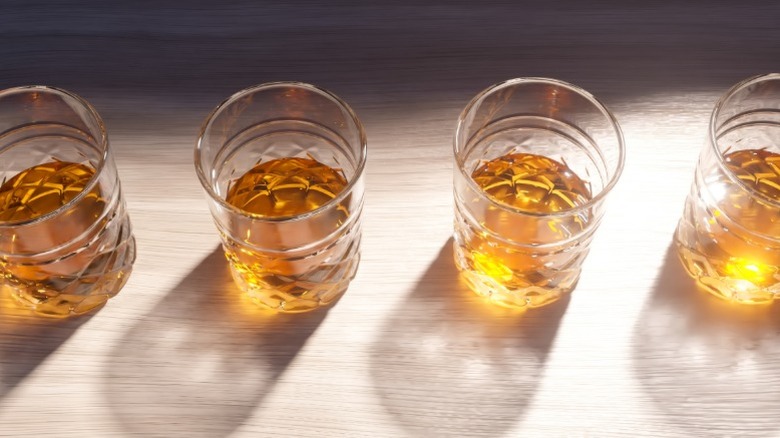 Glasses of whiskey poured neat lined up on a wood surface with sunlight shining on them.