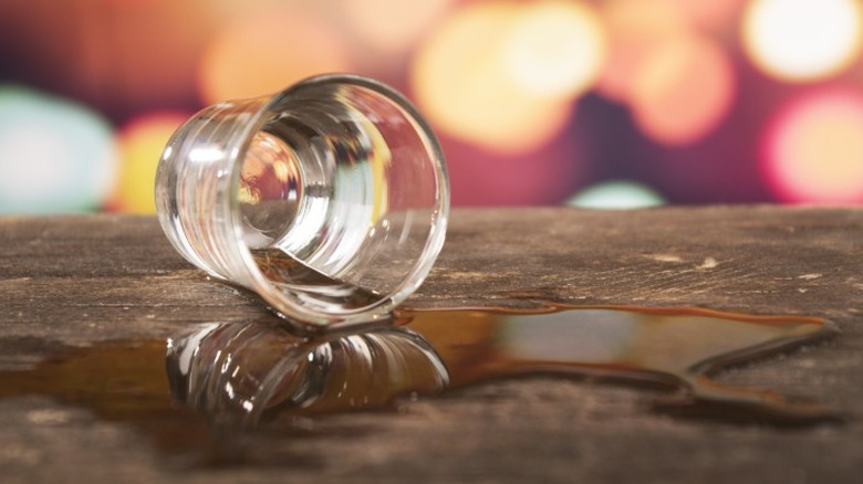 A shot glass of whiskey spilled on a wood surface.