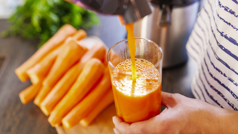Close-up of a person holding a glass of fresh carrot juice under a working juicer with a pile of carrots in the background