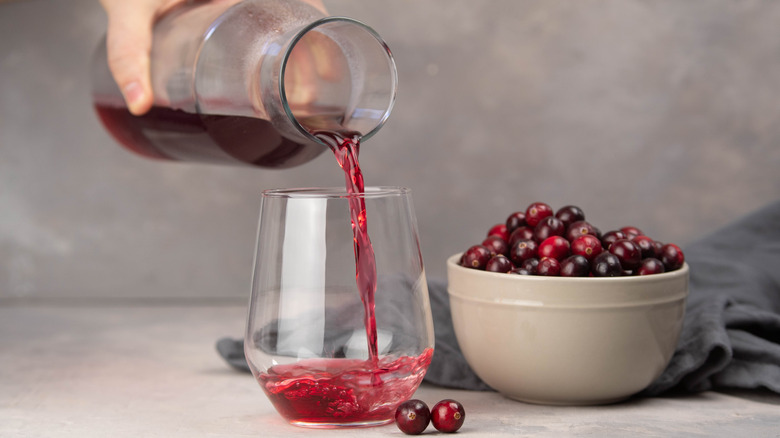 Close-up of a person pouring cranberry juice from a glass carafe into a juice glass, next to a bowl of cranberries
