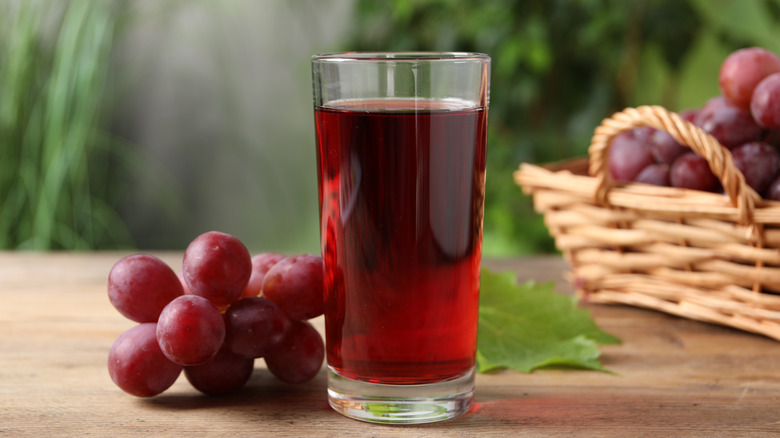 A glass of grape juice next to a bunch of grapes, with a grape leaf and a woven basket full of grapes in the background