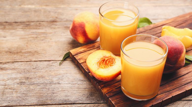 Two glasses of peach juice next to halved peaches on a wooden cutting board