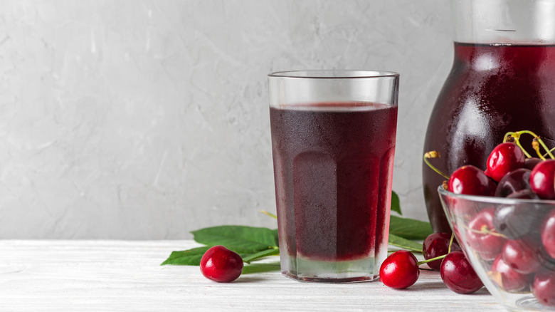 A glass of cherry juice next to cherry leaves, a glass bowl of cherries, and a pitcher of cherry juice