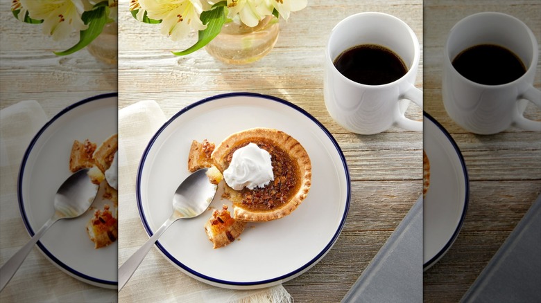 Freshness Guaranteed pecan pie with whipped cream on a navy blue-rimmed white plate next to a white mug of coffee and a vase with lillies