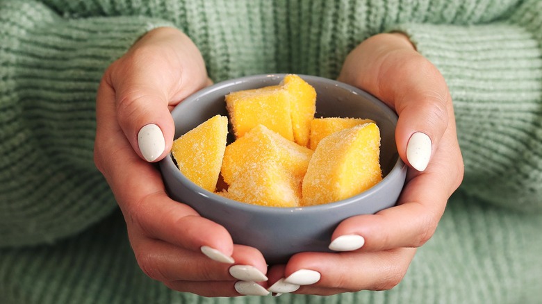 Two hands with white painted nails holding a gray bowl full of frozen mango chunks
