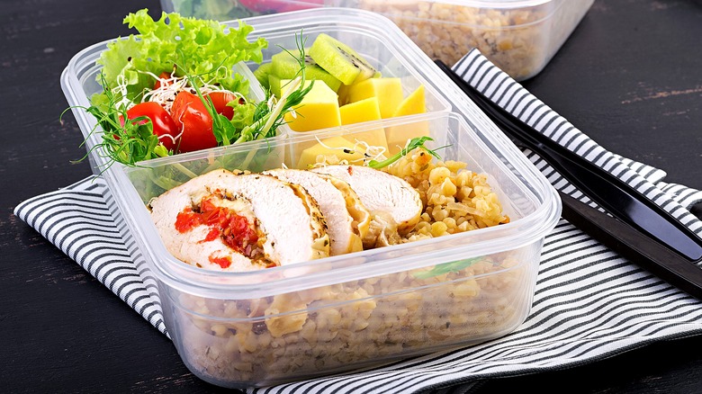 A clear, bento-style lunch box with sliced rotisserie chicken over cooked grains, green salad, and chopped fruit.