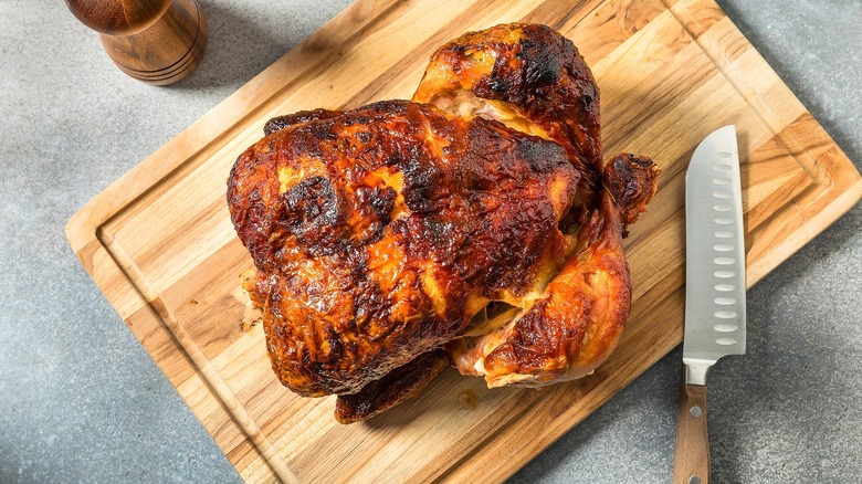 Whole rotisserie chicken on a wooden cutting board next to a sharp knife with a wooden handle.