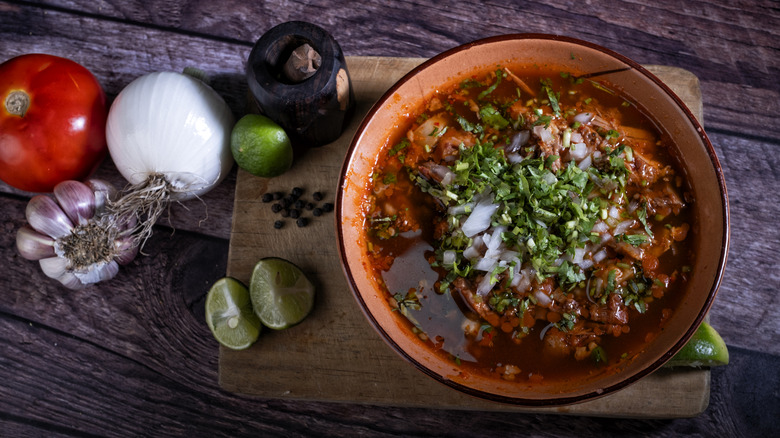 A bowl of birria with ingredients on a wooden cutting board