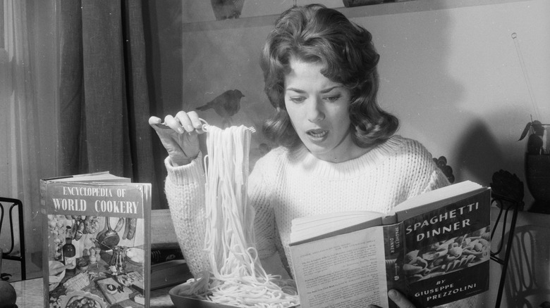 Black and white photo of a woman eating spaghetti and reading a cookbook